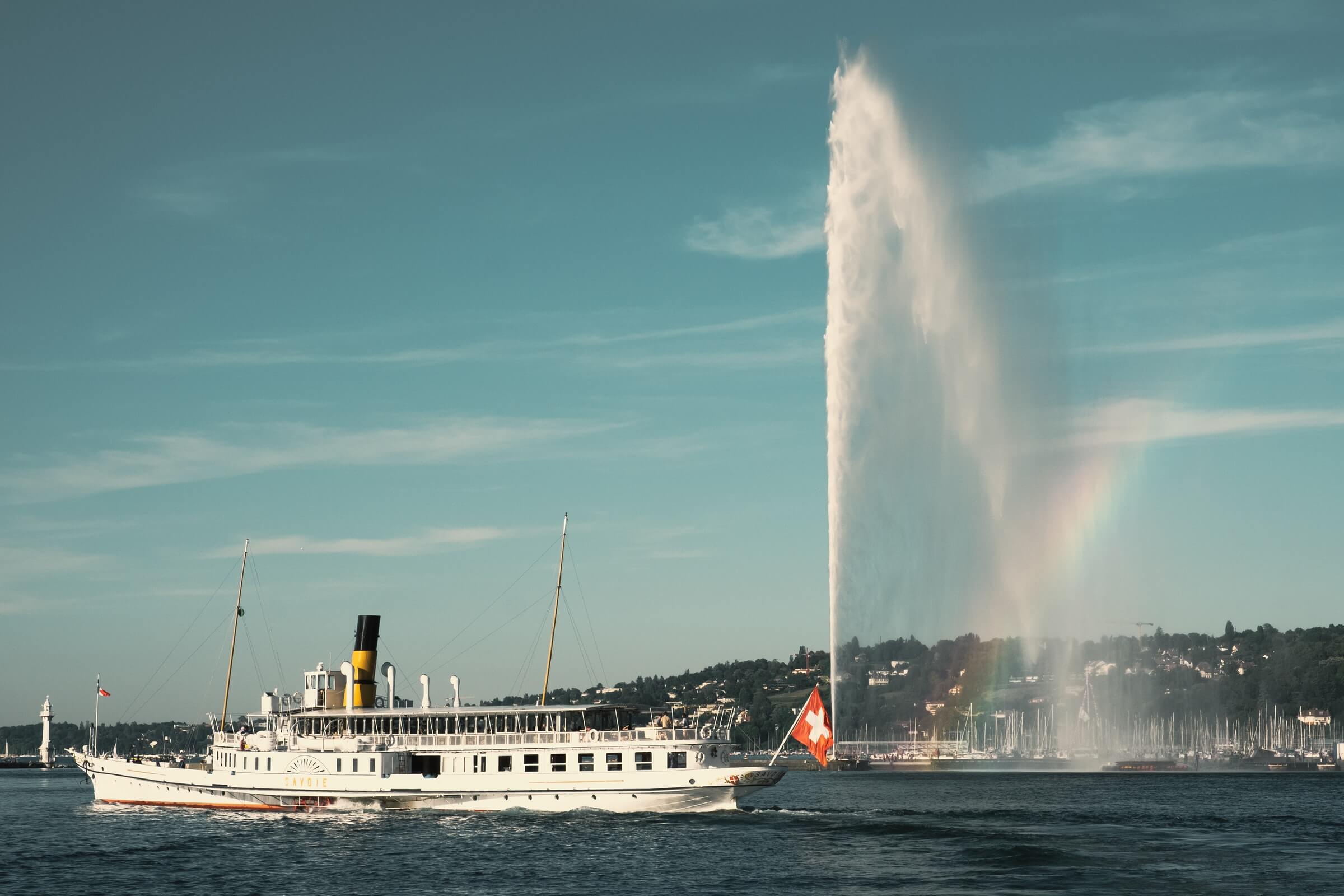Boat flying the Swiss flag on Lac Leman, Geneva, Switzerland with the Jet d'Eau and a rainbow in the background.
